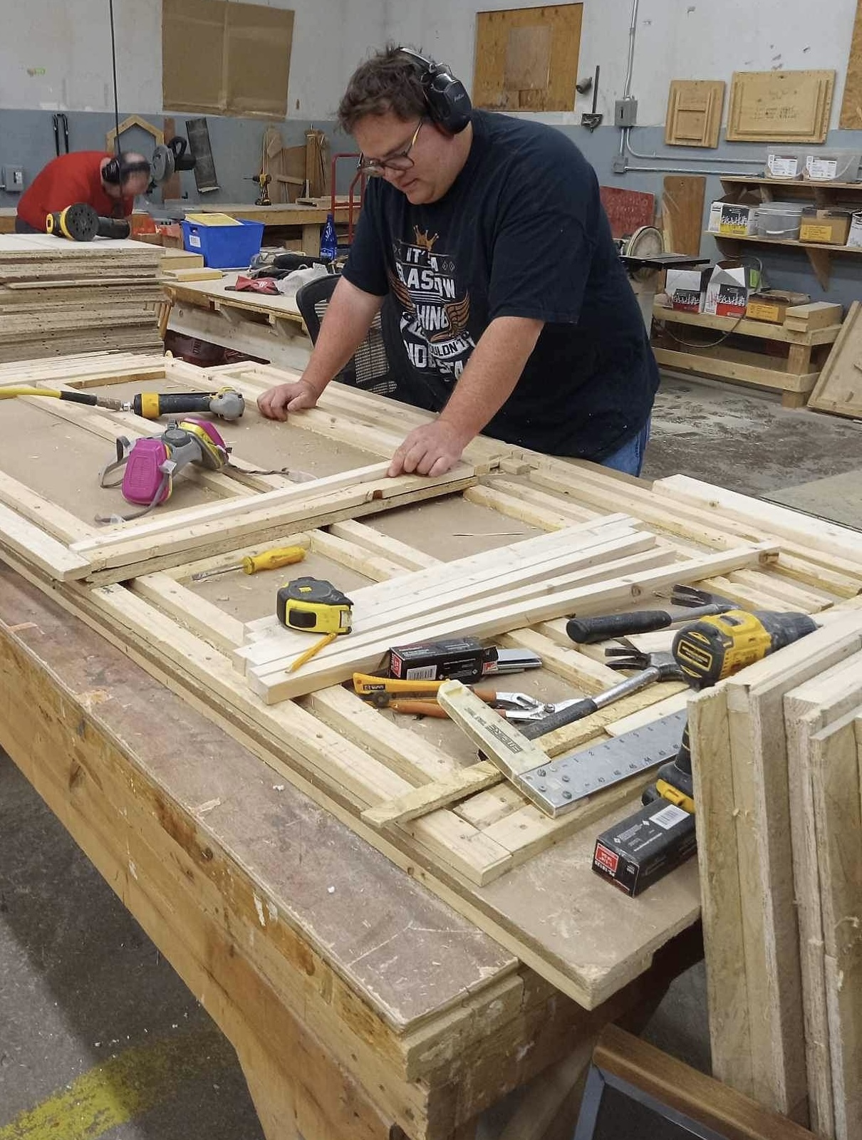 Wood shop employee, Alex, wearing ear protection, standing behind a work bench with a project in progress. Alex is focused on his project, not looking at the camera.