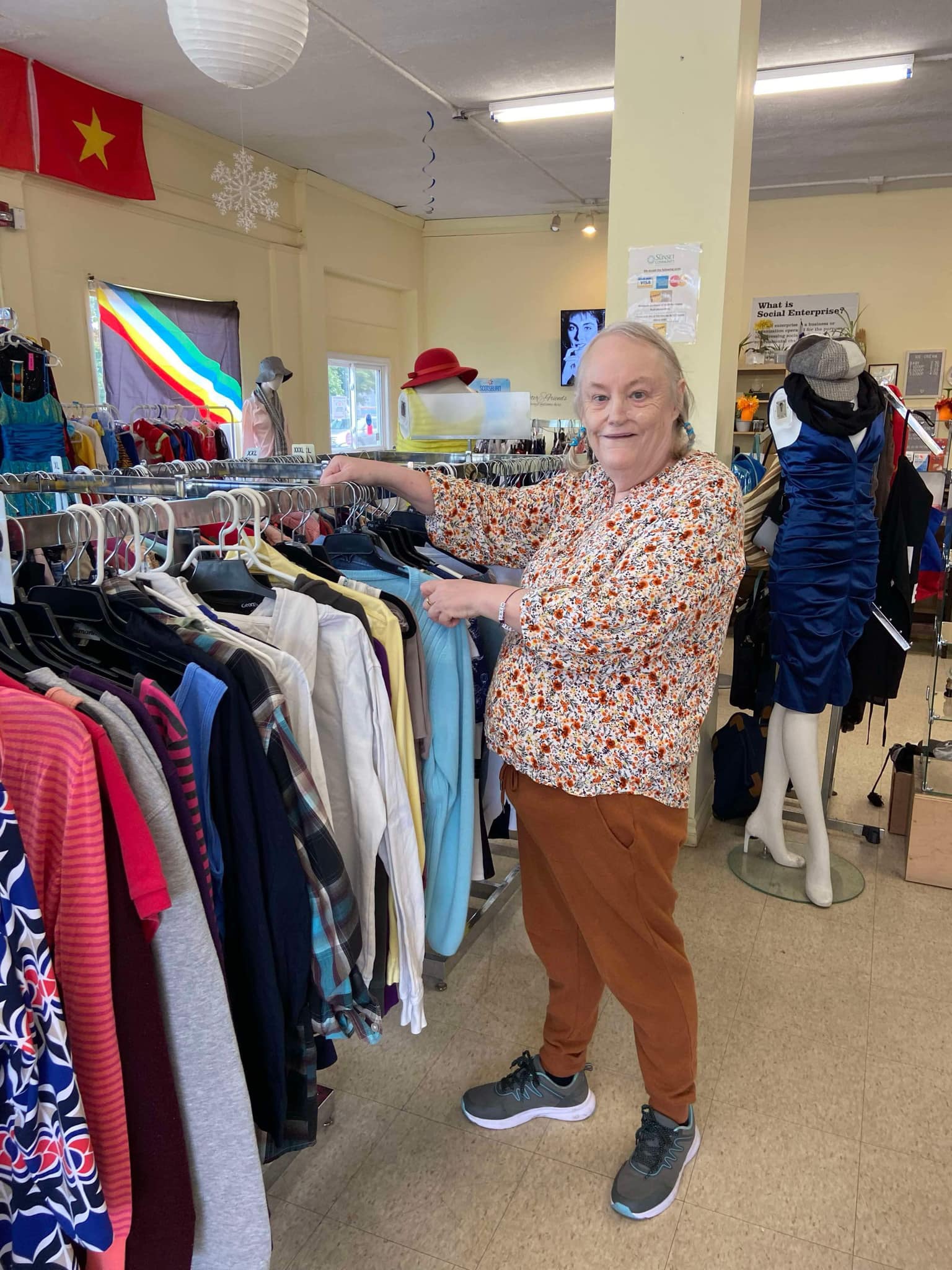 Thrift store employee, Theresa, standing next to a rack of women's tops, smiling at the camera.