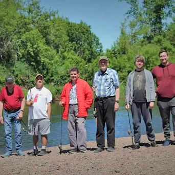 Program staff and participants posing with their fishing rods in front of a river. There are lush, green trees behind them.