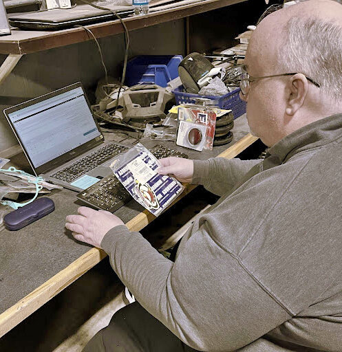 Crossroads employee, Mike, sits working at a computer, holding a part that he is inputting into a database.