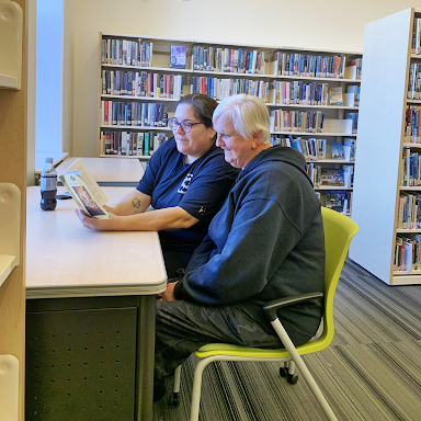 Program staff, Paula, and participant, Audrey, reading a book together, sitting at a table with a bookshelf behind them.