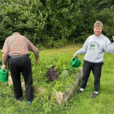 Stephen and David water a raised bed garden with green watering cans. David is facing away from the camera, Stephen waves and smiles at the camera.