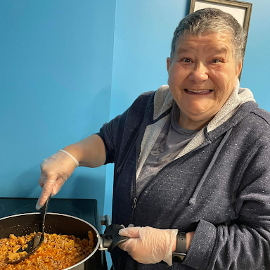 Cooking skills participant, Laura, smiles at the camera while stirring a pot of food on the stove.