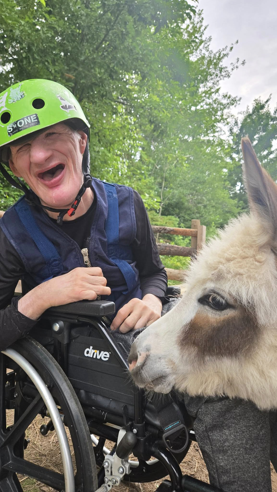 A person served at The Sunset Community smiles while visiting with a donkey outdoors. The person is seated in a wheelchair and wearing a bright green helmet surrounded by trees and nature.